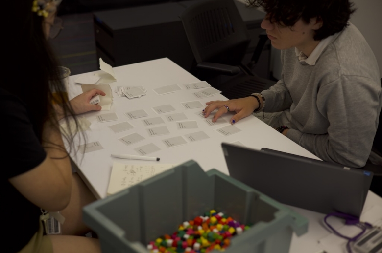 Two Tisch Summer High School Game Design students sit at a table with game cards arranged in rows. A box of colorful game pieces sits on the table, off to the side.